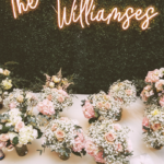 the williamses wedding neon sign with last name on greenery backdrop, flowers on table