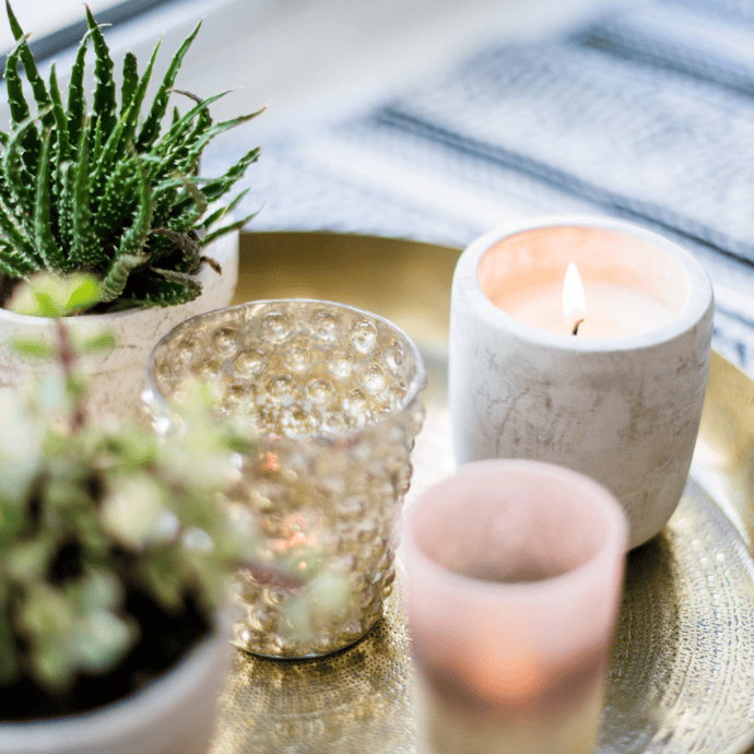 A decorative arrangement on a tray featuring a small potted succulent, a lit candle in a marble holder, a textured glass candle holder, and an out-of-focus plant in the foreground, placed on a woven fabric surface.