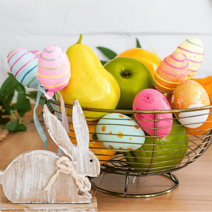 A wire basket filled with colorful Easter eggs and various fruits like apples and pears sits on a wooden table. A decorative wooden bunny with a twine bow is placed in front of the basket, adding to the festive scene.