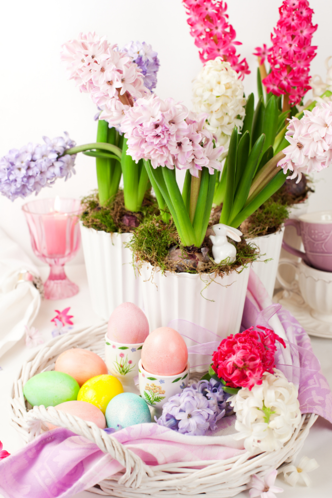 A spring-themed setting with blooming pink, purple, and white hyacinths in white pots. A small white rabbit figure hides among the flowers. Colorful Easter eggs sit in a woven basket with a pink cloth. A pink candle and cup are in the background.