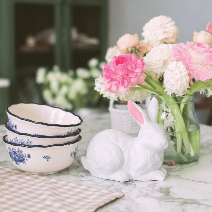 A marble table presents three stacked white bowls with blue floral designs, a white ceramic rabbit figurine, and a vase of pink and white peonies—perfect Easter decor. A beige checkered cloth rests nearby, while the soft-focus background reveals kitchen cabinets.