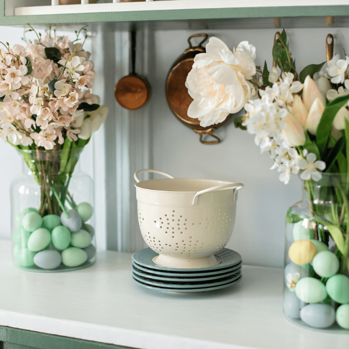 A kitchen counter adorned with Easter Kitchen Decor showcases two glass vases filled with pastel-colored eggs and white flowers. At the center, a white colander rests on blue plates, while copper pans hang in the background, adding a rustic touch of cooking magic.