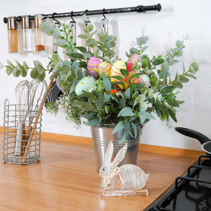 A kitchen counter features a metal vase with greenery and vibrant Easter egg decorations, exuding charming Easter Kitchen Decor. Nearby, a decorative wooden rabbit adds whimsy. In the backdrop, a utensil holder with whisks and a spice rack hints at Cooking Magic ready to unfold.