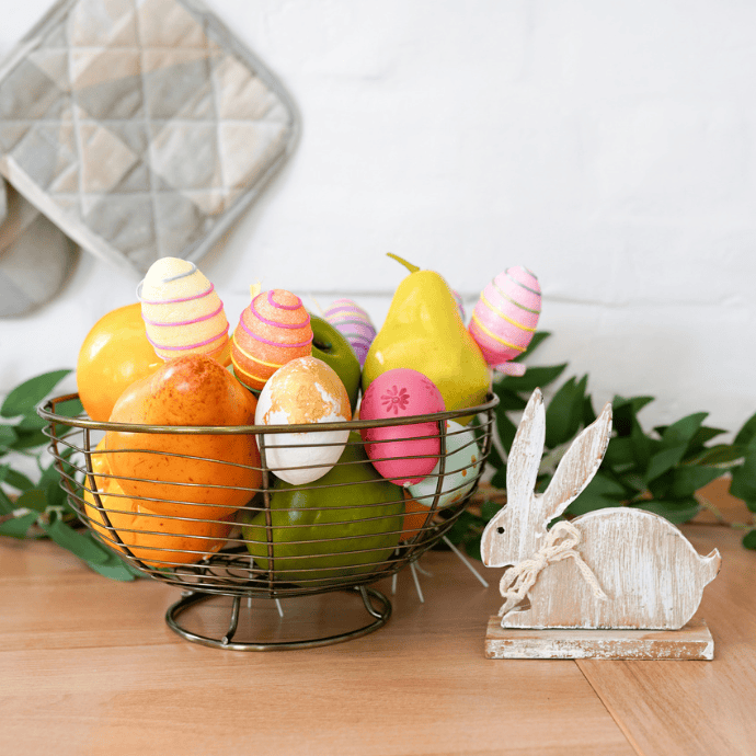 A wire basket filled with pears and decorated eggs graces the wooden table, embodying the charm of Easter Kitchen Decor. Nearby, a white wooden rabbit figurine sits, while a quilted potholder hangs on the white brick wall, green leaves peeking through like a touch of Cooking Magic.