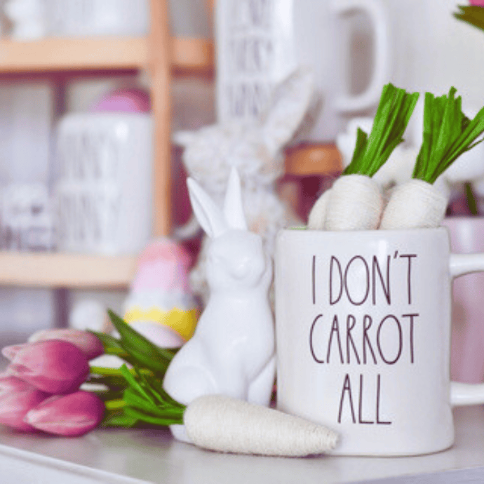 White mug with "I DON'T CARROT ALL" text, holding decorative white carrots with green tops. Beside the mug is a white rabbit figure and pink tulips, perfect for Easter kitchen decor. The background features a soft-focus interior filled with more decorative items.