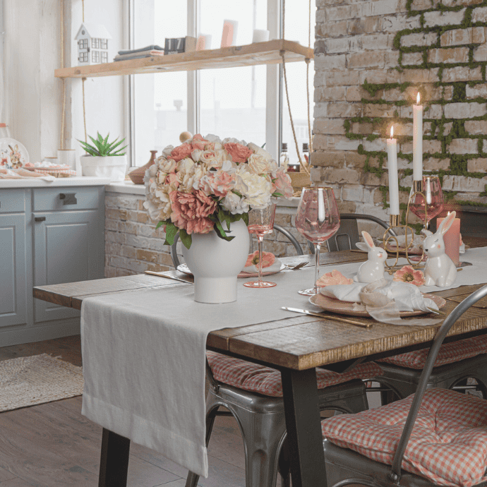 A rustic dining room with a wooden table set for a meal is adorned with enchanting Easter kitchen decor. Pink and white flowers in a vase, bunny-shaped accents, pink wine glasses, and lit candles add charm against the backdrop of a brick wall and white kitchen cabinets.
