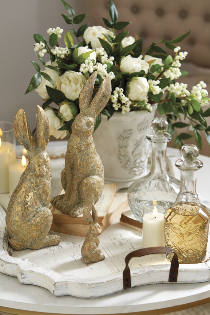 A decorative setup featuring three rabbit figurines of varying sizes in earthy tones on a white tray for a perfect Easter touch. Nearby, glass decanters hold amber liquid. A lit candle and a floral arrangement with white roses and greenery complement the cozy living room decor.