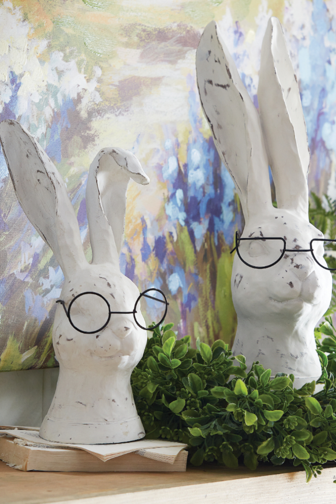 Two whimsical white rabbit busts with black round glasses sit on a wooden shelf. Each rabbit has a slightly weathered texture and long ears. Surrounded by green foliage and set against a colorful floral backdrop, they offer charming Easter decor ideas for a spring refresh.