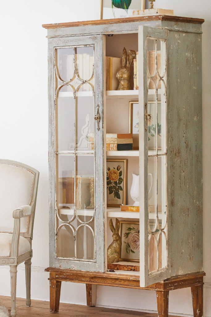 A vintage cabinet with glass doors stands open, displaying books, a framed botanical print, and decorative items like a golden rabbit figure. To the left, a cream-colored upholstered chair is partially visible. The setting is cozy and elegant.