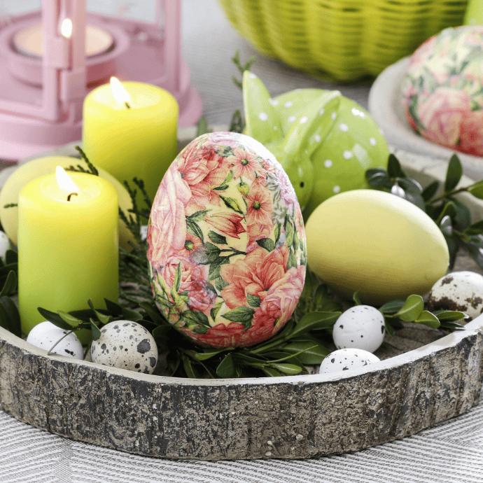 A decorative Easter arrangement brightens your living room decor with a large egg adorned in floral designs, surrounded by speckled eggs, yellow candles, and lush greenery on a wooden tray. A charming green polka-dotted bunny and pink lantern add to the spring refresh in the background.