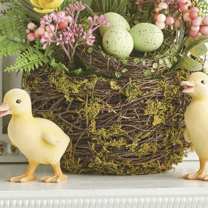 A decorative nest with green speckled eggs sits among pink and yellow flowers, capturing the essence of Easter living room decor. Nearby, two yellow duckling figurines rest on a white shelf, creating a charming spring-themed arrangement.