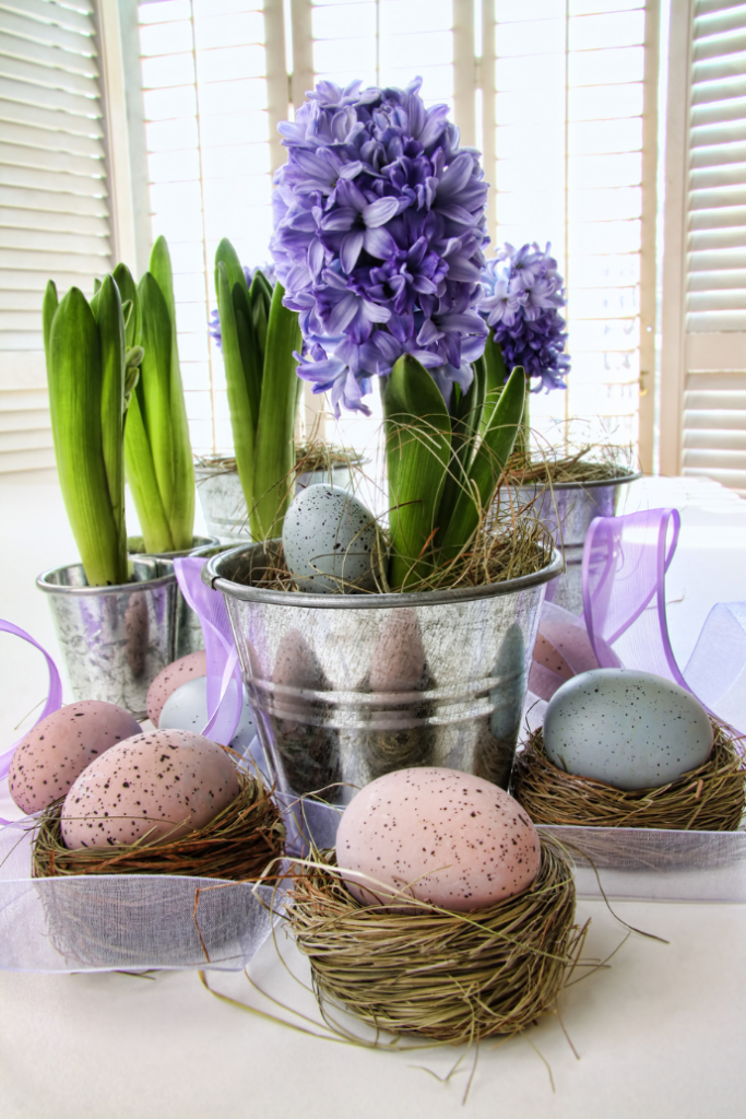 Metal buckets with purple hyacinths and green leaves brighten up the living room decor, capturing the essence of a spring refresh. Speckled pastel eggs, some blue and some pink, rest in nests of straw around the pots. Soft light filters through the window blinds in the background.