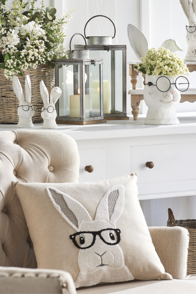 A cozy room featuring a beige armchair with a pillow depicting a rabbit wearing glasses. Embrace Easter living room decor with decorative rabbit figures, lit lanterns, and spring flowers in baskets on a white shelf.