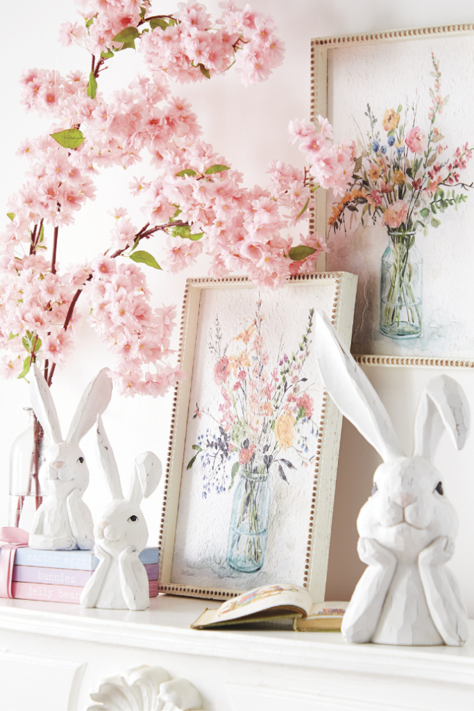 A mantel adorned with cherry blossom branches, framed floral paintings, and white ceramic rabbit figurines exudes a bright, spring refresh. Some rabbits perch on books while another rests beside an open book, creating perfect Easter living room decor.