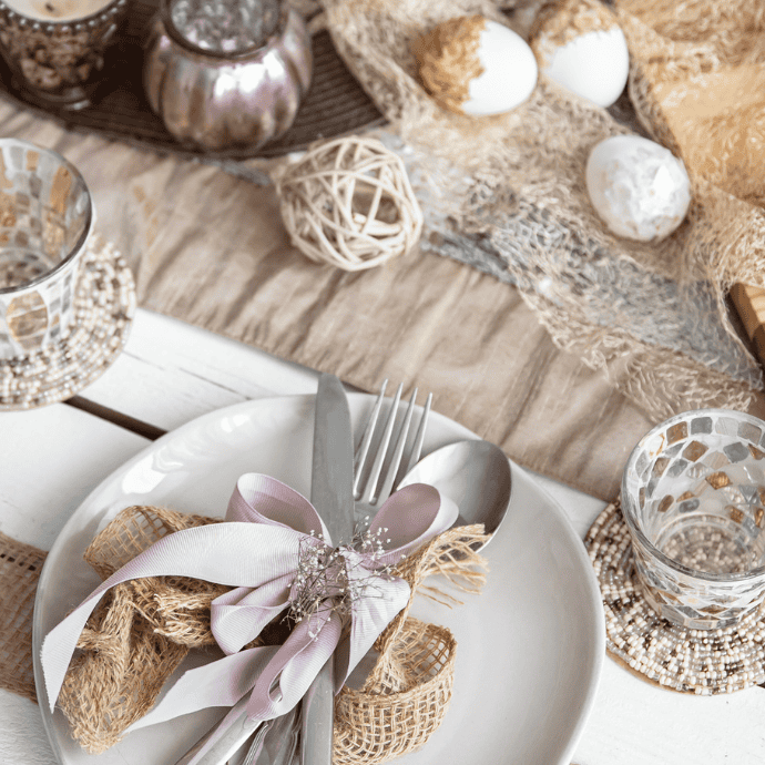 A white plate with a fork and knife wrapped in burlap and ribbon rests on a rustic table, embodying elegant Easter table settings. Decorative eggs, woven balls, and patterned glassware enhance the scene in a light brown and beige color palette.