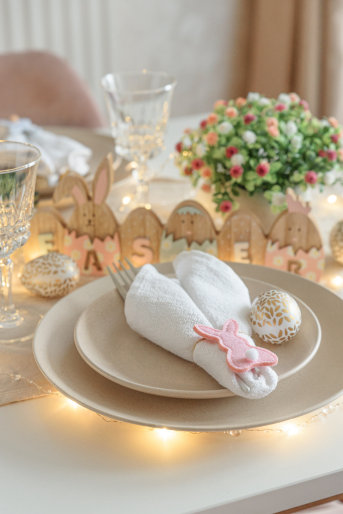 Experience elegant Easter table settings with a beige plate and silverware wrapped in a white napkin, secured by a charming pink bunny clip. Adorned with decorated eggs, crystal glasses, flowers, a wooden bunny "EASTER" sign, and twinkling string lights for a festive touch.