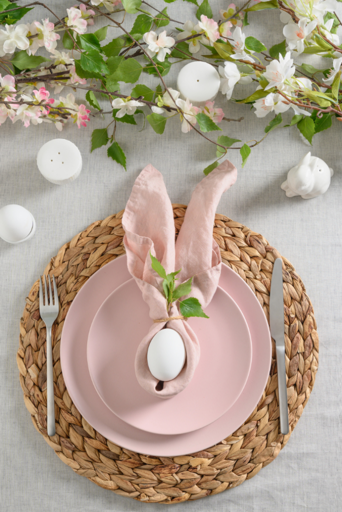 An elegant Easter table setting showcases a pink plate with a napkin folded like rabbit ears, cradling a white egg, all placed on a woven placemat. Beautiful flowers and candles surround the setting, creating an inviting spring-themed ambiance perfect for Easter table ideas.