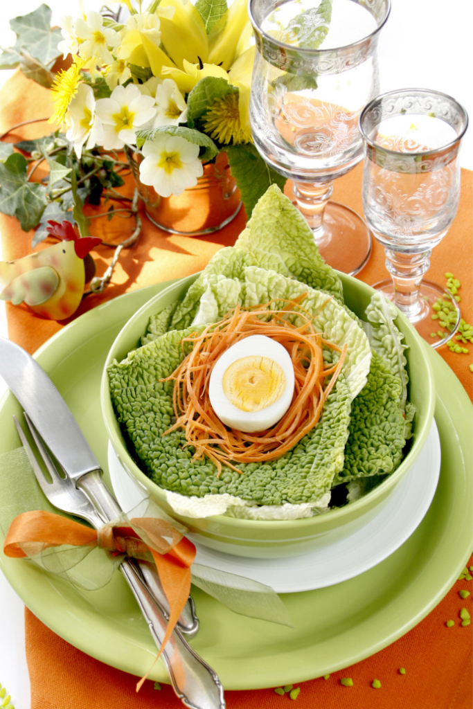 An elegant Easter table setting showcases a vibrant green salad bowl filled with crisp lettuce, shredded carrots, and half a boiled egg. It's set on an orange napkin. The arrangement includes two glasses, floral decorations, and a knife and fork elegantly tied with an orange ribbon.