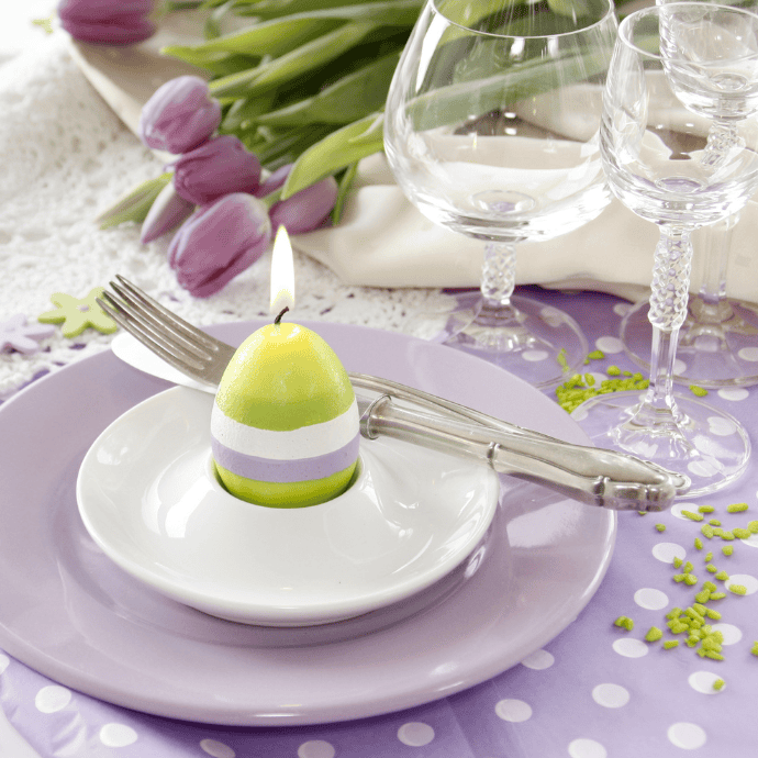 An elegant Easter table setting with a lilac tablecloth features a lit, striped egg-shaped candle in a white dish, topped with silver cutlery. Purple tulips and lace add a decorative touch, alongside elegant glassware, creating the perfect atmosphere for a beautiful meal.