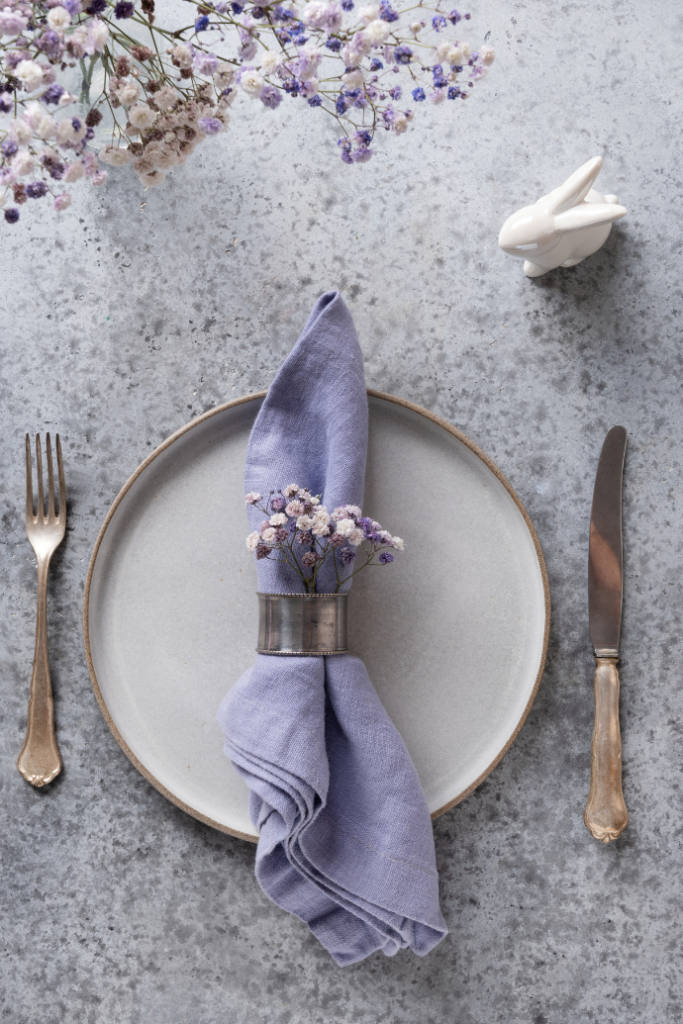 An elegant Easter table setting features a gray plate, lavender napkin adorned with white flowers, and a silver napkin ring. A silver fork and knife rest on the textured surface, with a small white rabbit ornament adding charm at the top.