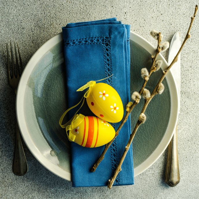 A table setting with a gray plate and green bowl, featuring a blue napkin topped with two decorated yellow Easter eggs. A fork and knife are on either side, and a sprig of pussy willow rests on the plate.