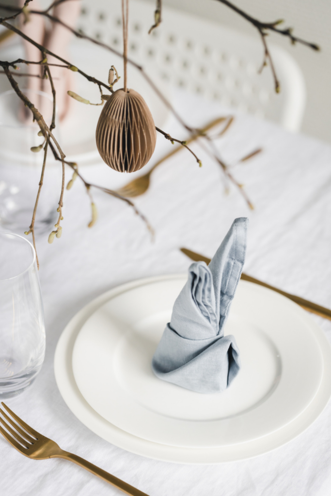 A table is set with a white plate and a light blue, folded napkin. A glass sits nearby, along with a gold fork and knife. Above, a decorative brown paper egg hangs from a twig. The tablecloth is white.
