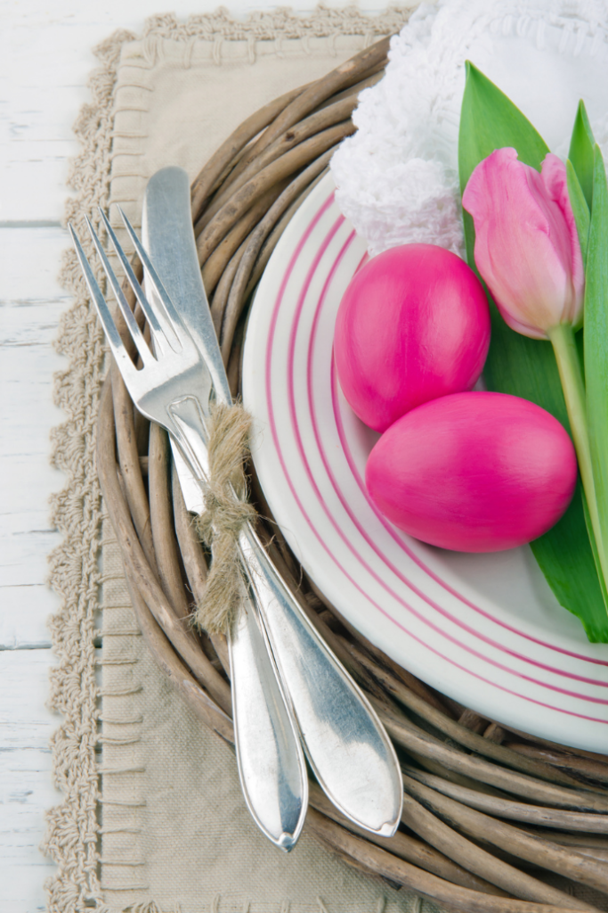A plate with two pink eggs and a pink tulip rests on a wicker charger. Silverware tied with twine is placed beside it on a fringed cloth. The setting is on a white wooden table.