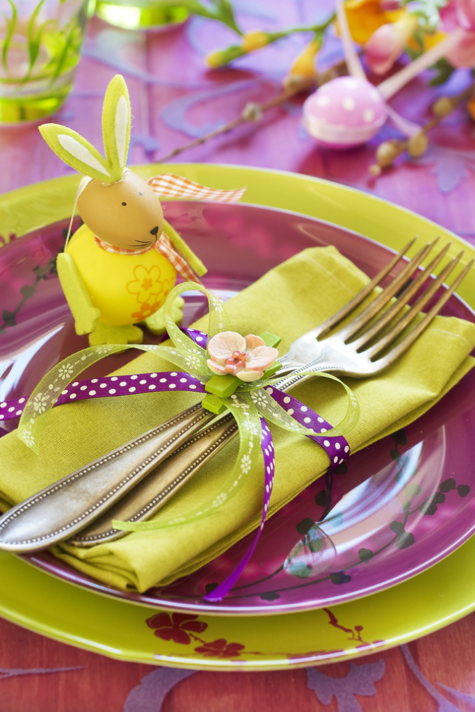 A vibrant Easter table setting features a green plate, a purple glass plate, and a lime green napkin adorned with a flower and ribbon. Silverware is placed on the napkin, and a yellow bunny decoration sits beside it.