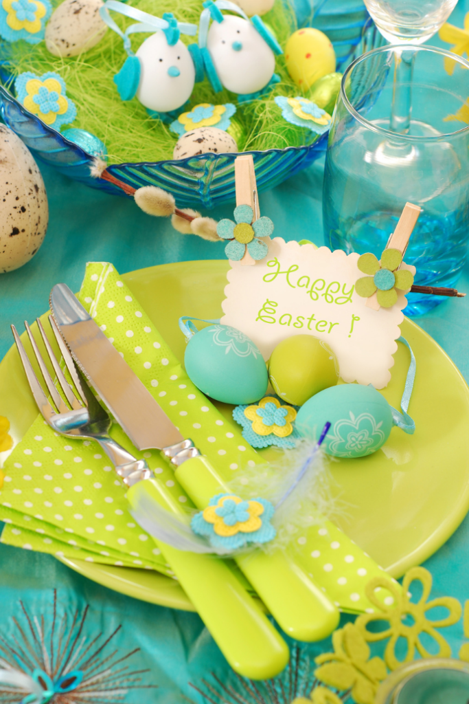 A festive Easter table setting with a yellow plate, green cutlery, and polka dot napkin. Decorated eggs, including a "Happy Easter" note, are displayed. Blue and yellow tones dominate, with bird decorations and green grass accents.