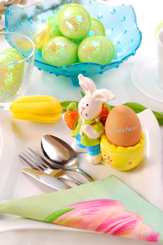 A festive Easter table setting with a ceramic bunny holding a carrot next to a yellow egg cup with an egg labeled "Easter." Nearby are cutlery, a yellow tulip, and a bowl of decorated eggs. A green floral napkin accents the arrangement.