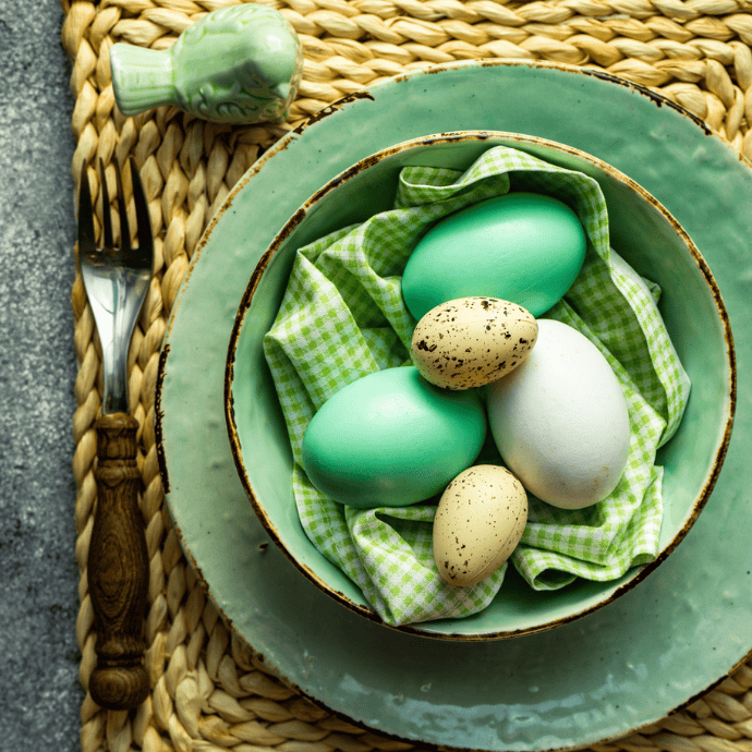 A mint green plate holds dyed eggs in shades of green and white, placed on a green checkered cloth. The setting includes a woven placemat, a fork on the left, and a ceramic bird in the background.