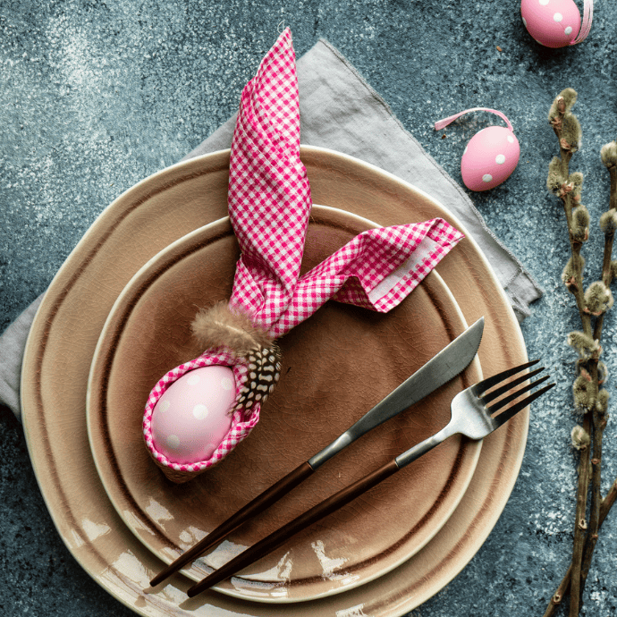 A festive Easter table setting with a pink polka dot egg wrapped in a checkered napkin resembling bunny ears. The plate is layered with neutral-colored linens, accompanied by a fork and knife. Pussy willow branches are on the side, adding a spring touch.