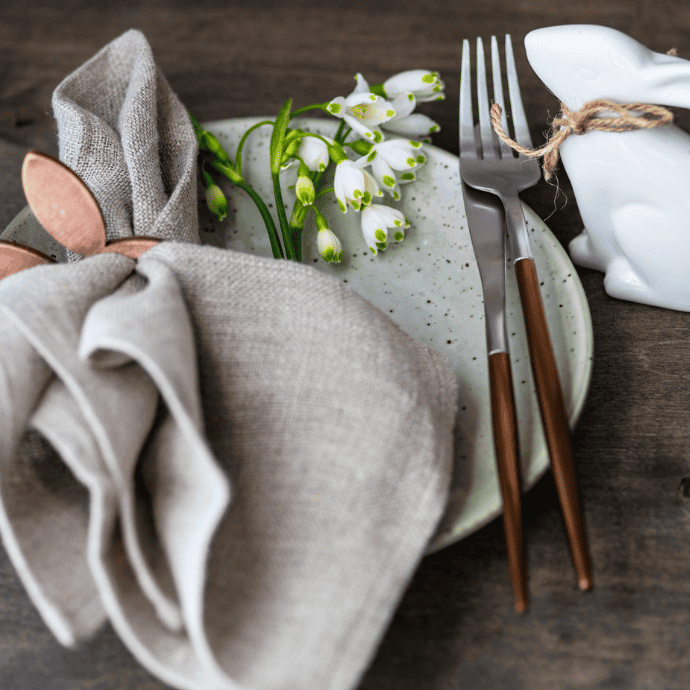 A table setting featuring a speckled plate with a folded linen napkin and wooden napkin ring shaped like bunny ears. Snowdrop flowers, a fork and knife, and a ceramic rabbit tied with twine complete the decor on a dark wooden table.