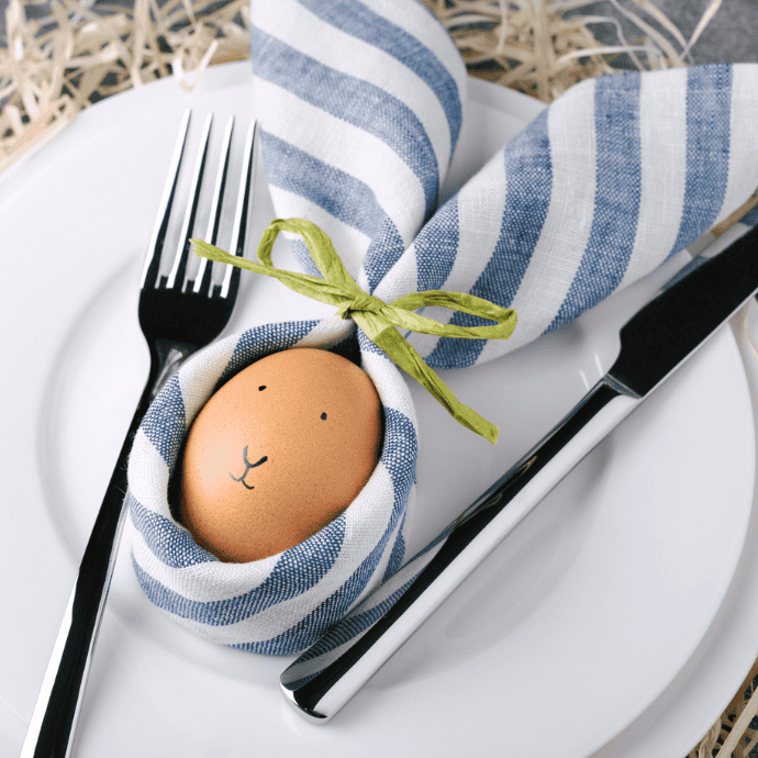 A brown egg with a drawn face is wrapped in a striped blue and white cloth, resembling rabbit ears, and tied with a green bow. It is placed on a white plate with a fork and knife on either side.
