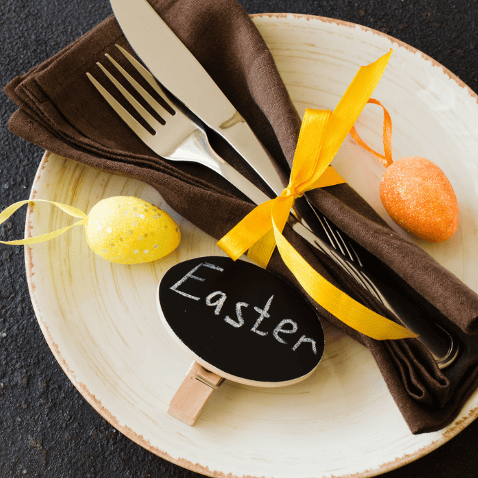 A table setting includes a beige plate with a brown napkin tied with a yellow ribbon, holding a knife and fork. Two decorative egg ornaments, one yellow and one orange, are nearby. A small chalkboard sign with "Easter" written on it rests on the plate.