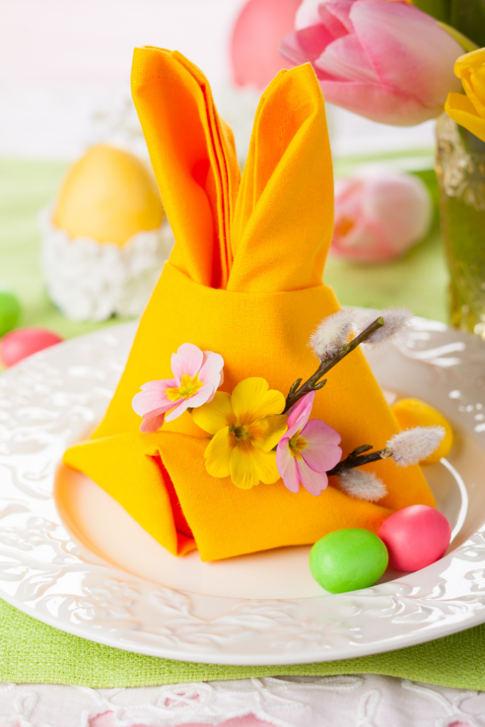 A decorative table setting with a yellow napkin folded into bunny ears on a white plate. The napkin is adorned with small flowers. Colorful Easter eggs are scattered around, and pink tulips are in the background.