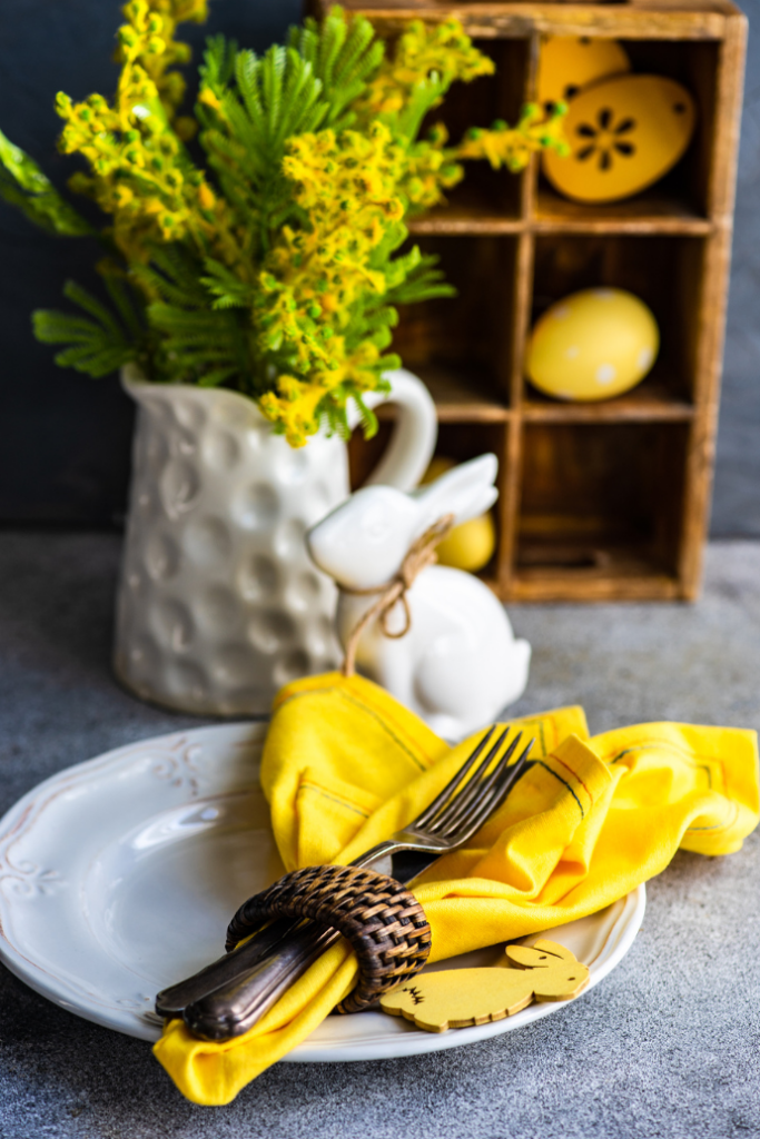 A table setting with a white plate, yellow napkin, and cutlery wrapped with a dark woven ring. A decorative white rabbit and vase of yellow flowers are in the background. A wooden shelf holds yellow ornaments.
