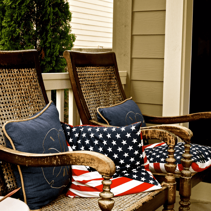 Two wooden chairs with woven seats and backs sit on a porch, each adorned with blue pillows and patriotic porch decor—cushions featuring the American flag's stars and stripes. A white railing and green bush are in the background.