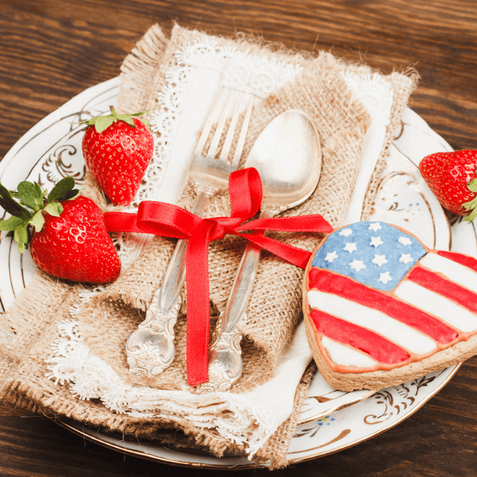 A plate with a burlap napkin holding a fork and spoon tied with a red ribbon, surrounded by fresh strawberries and a heart-shaped cookie decorated with the American flag—perfect patriotic table decor.