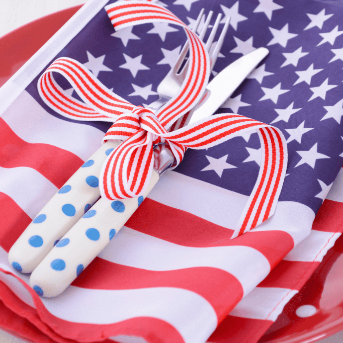 A fork and knife with white handles covered in blue polka dots are tied with a red-striped ribbon, placed on a napkin resembling the American flag atop a red plate, creating festive patriotic table decor.