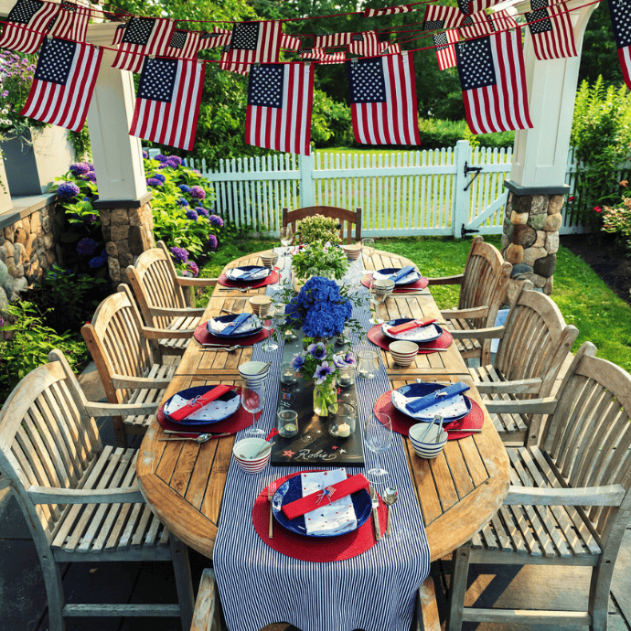 A wooden outdoor dining table features patriotic table decor with red, white, and blue accents, American flags, striped napkins, and flowers. Wooden chairs encircle the scene under a pergola adorned with hanging flags in a charming garden setting.