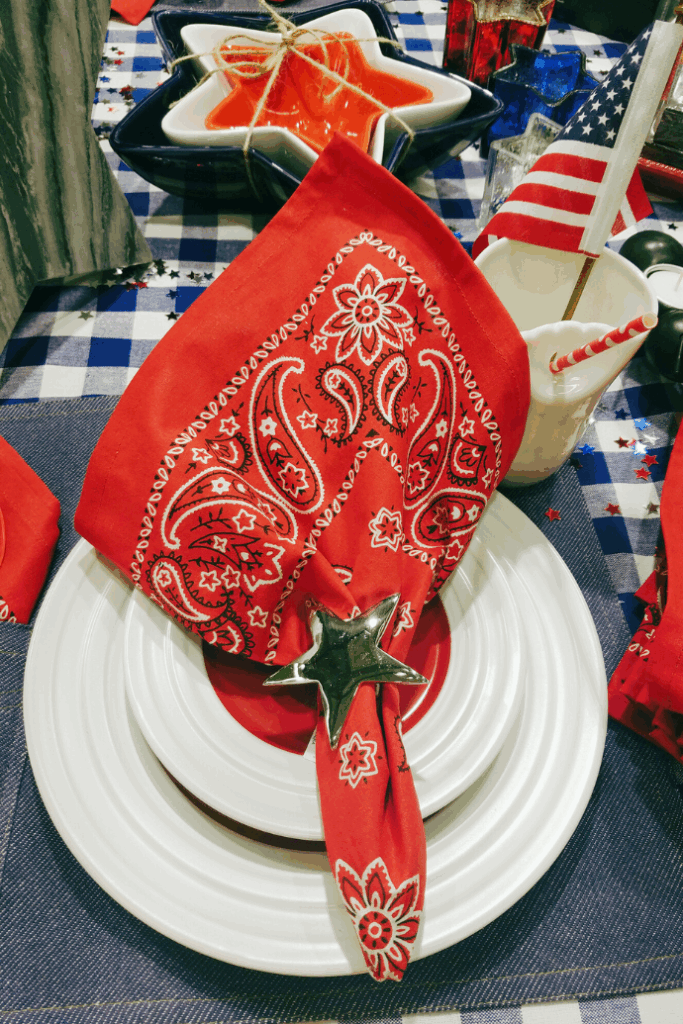 A patriotic table decor scene features a red bandana napkin in a star-shaped napkin ring on a white plate. An American flag and star-shaped dish add charm to the display, all set atop a blue checkered tablecloth.