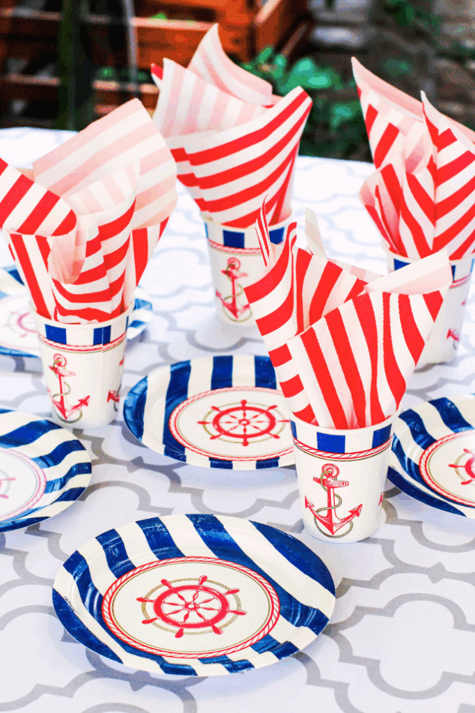 Nautical-themed party table with patriotic table decor features plates and cups adorned with anchors and ship wheels, red and blue stripes, and red-striped napkins arranged in cups on a white patterned tablecloth.