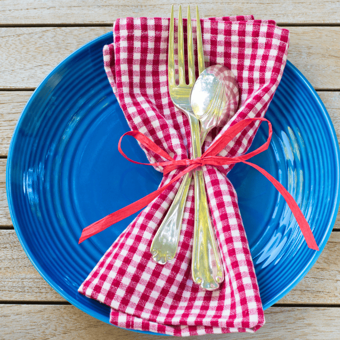 A blue plate holds a red and white checkered napkin tied with a red ribbon, with a fork and spoon on top—perfect patriotic table decor set on a wooden table.