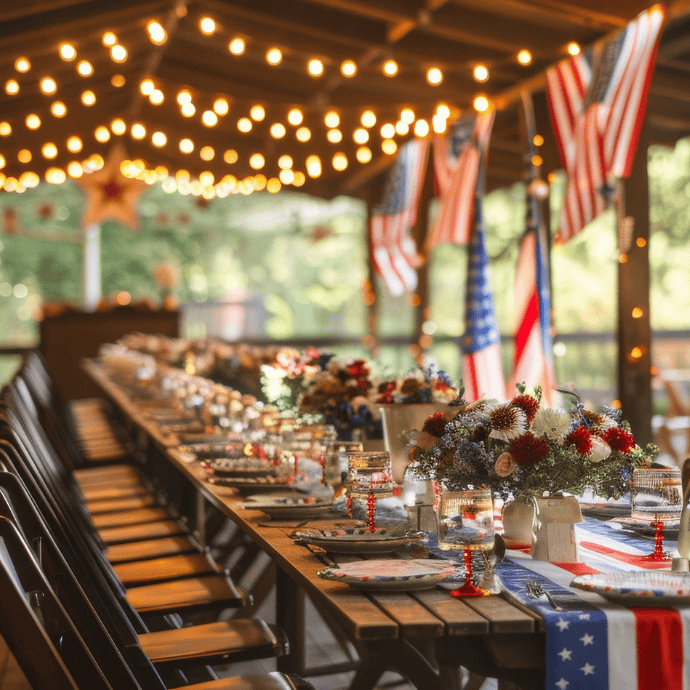 A long outdoor table features patriotic table decor with American flags, red, white, and blue flowers, and string lights overhead, all set for a festive gathering under a covered patio.