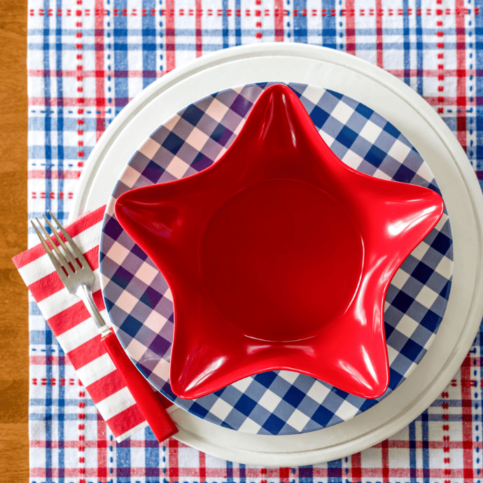 A red star-shaped bowl sits on a blue and white checkered plate, with a white plate beneath—patriotic table decor accented by a fork and striped red and white napkin, all atop a red, white, and blue plaid tablecloth.