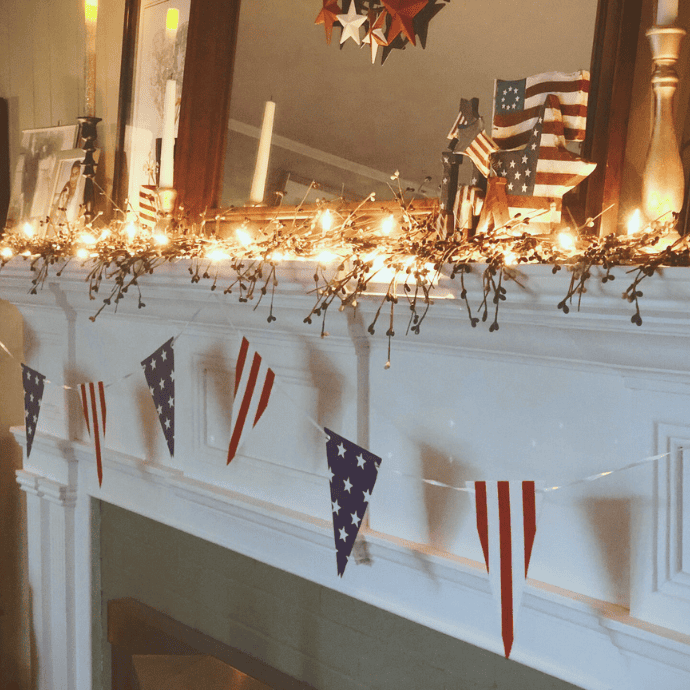 A white mantel decorated with string lights, American flag-themed bunting, small American flags, and patriotic star ornaments, creating a festive, patriotic display.