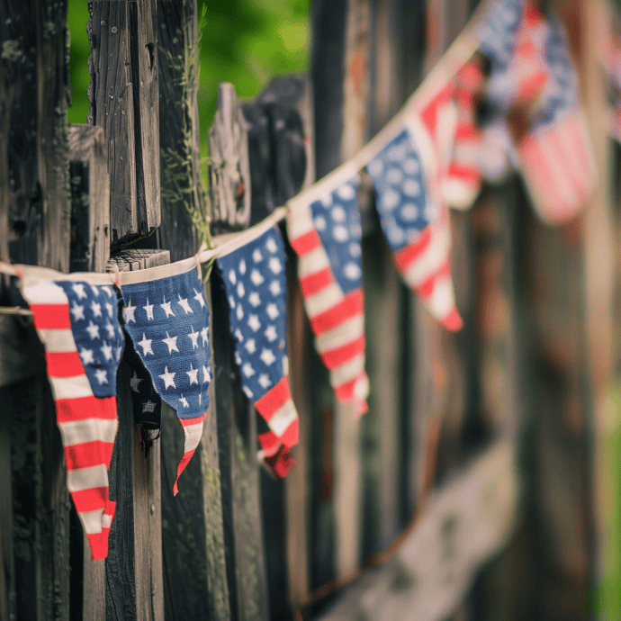 Weathered American flag bunting hangs along a rustic wooden fence, with the stars and stripes pattern of the fabric slightly faded. The background is softly blurred, giving a warm, nostalgic feel.