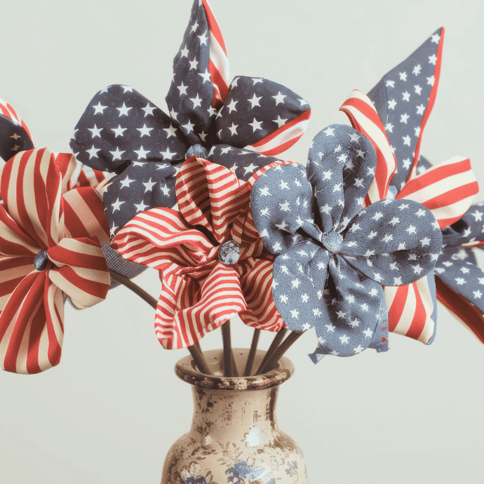 A ceramic vase holds fabric flowers made from American flag-patterned material, featuring red and white stripes and blue with white stars. The arrangement is set against a plain light background.