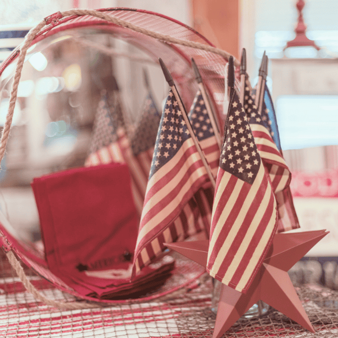 Small American flags are arranged in a rustic star-shaped holder next to red napkins and a decorative wire basket on a plaid tablecloth, creating a patriotic display.